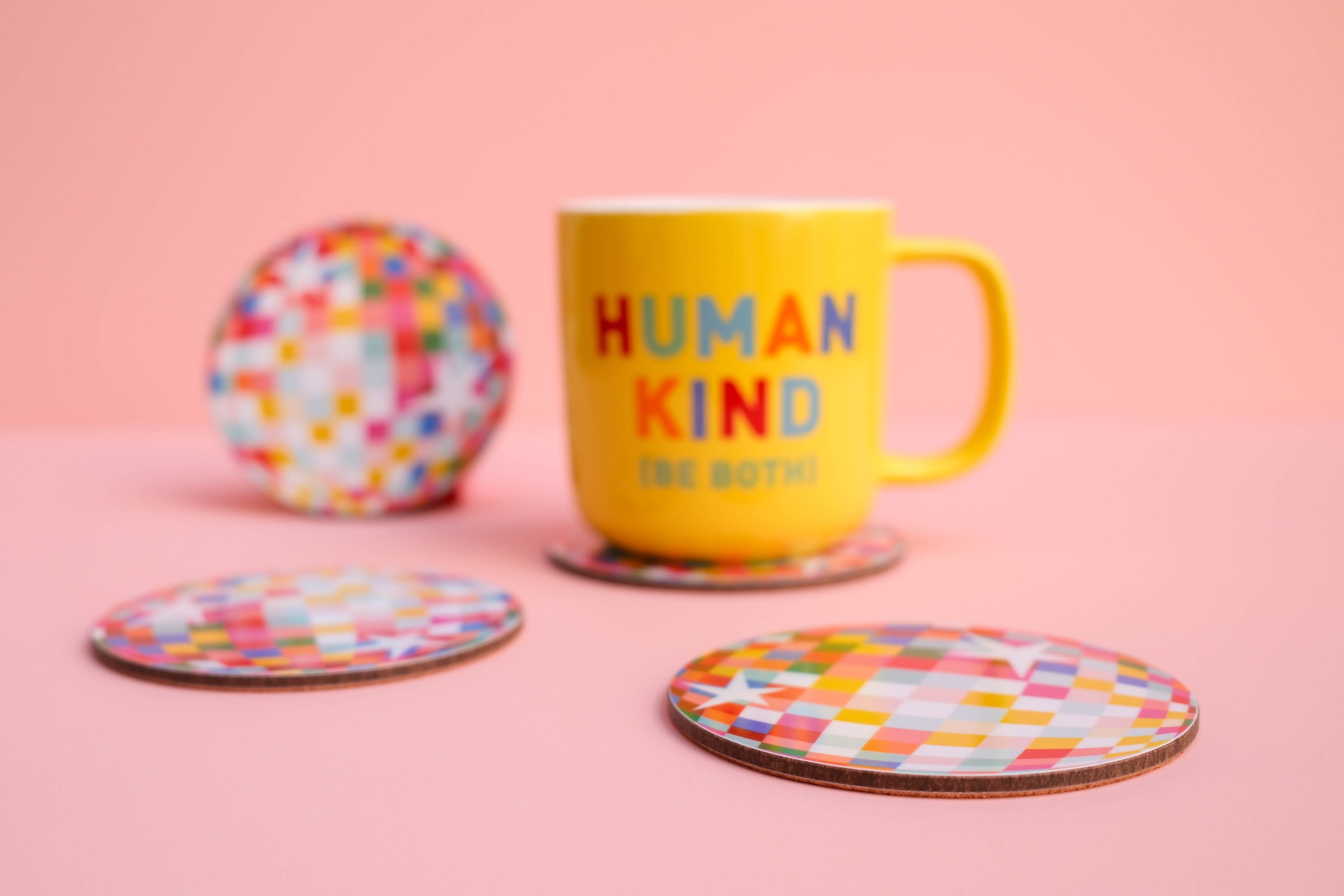 Yellow mug with 'Human Kind The Both' text on a pink background with colorful coasters.