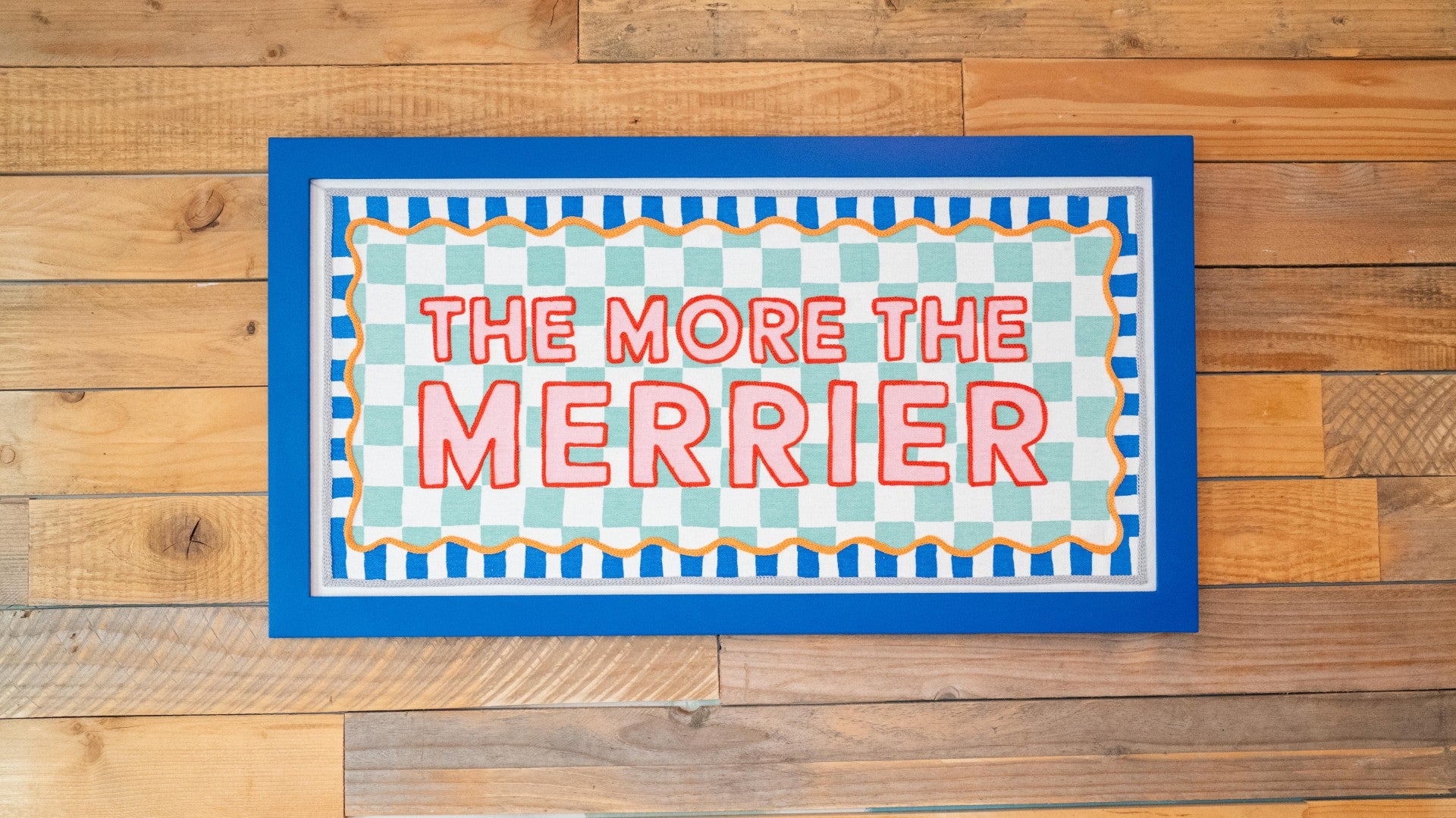 Decorative fabric sign  with 'The More The Merrier' text on pallet wall