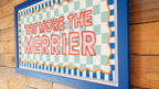 Decorative sign with 'The More The Merrier' text on a checkered background