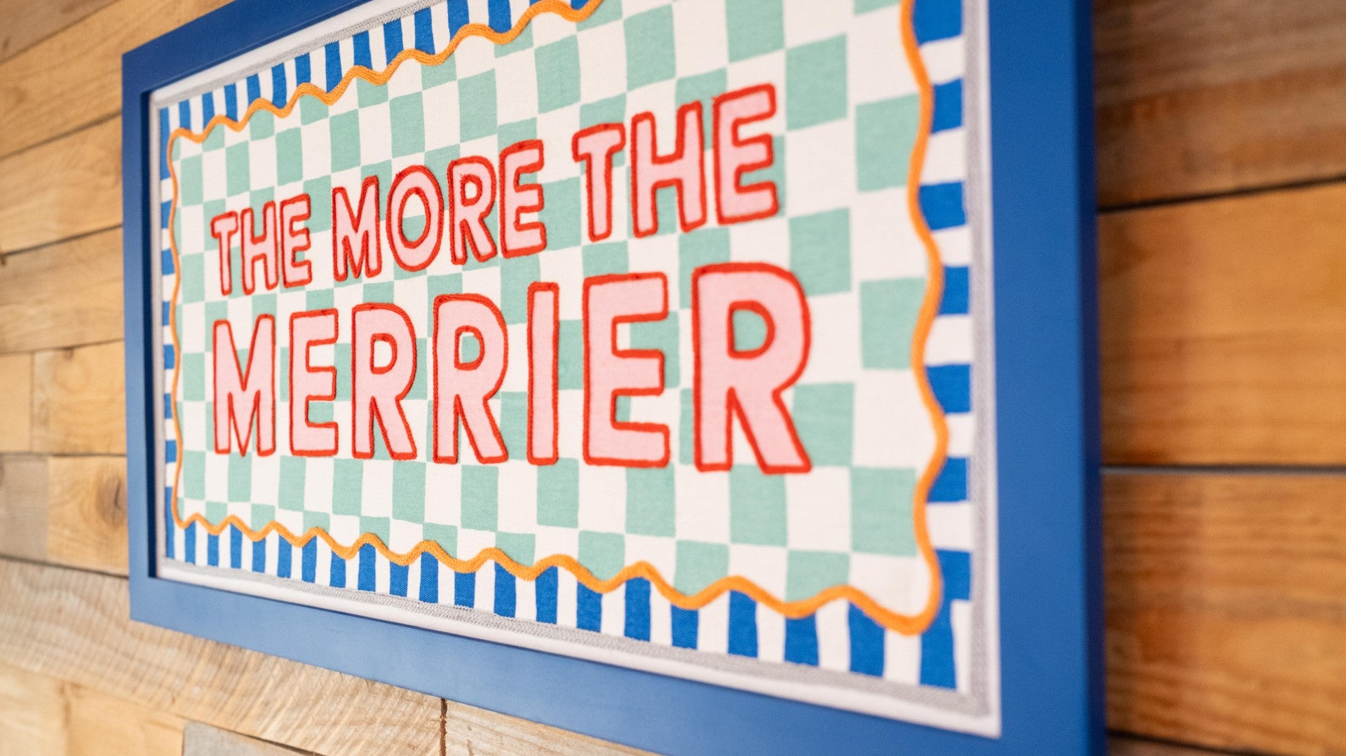 Decorative sign with 'The More The Merrier' text on a checkered background
