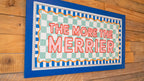 Decorative sign with 'The More the Merrier' text on a wooden background