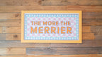 Decorative sign with 'The More The Merrier' text on a wooden background