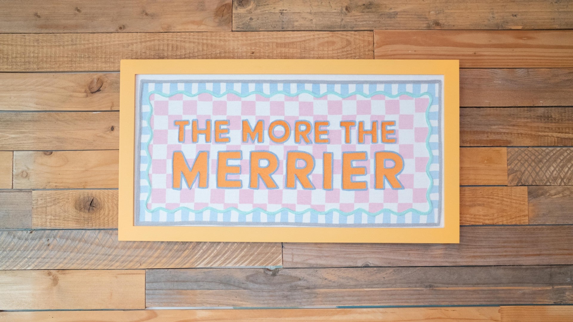 Decorative sign with 'The More The Merrier' text on a wooden background