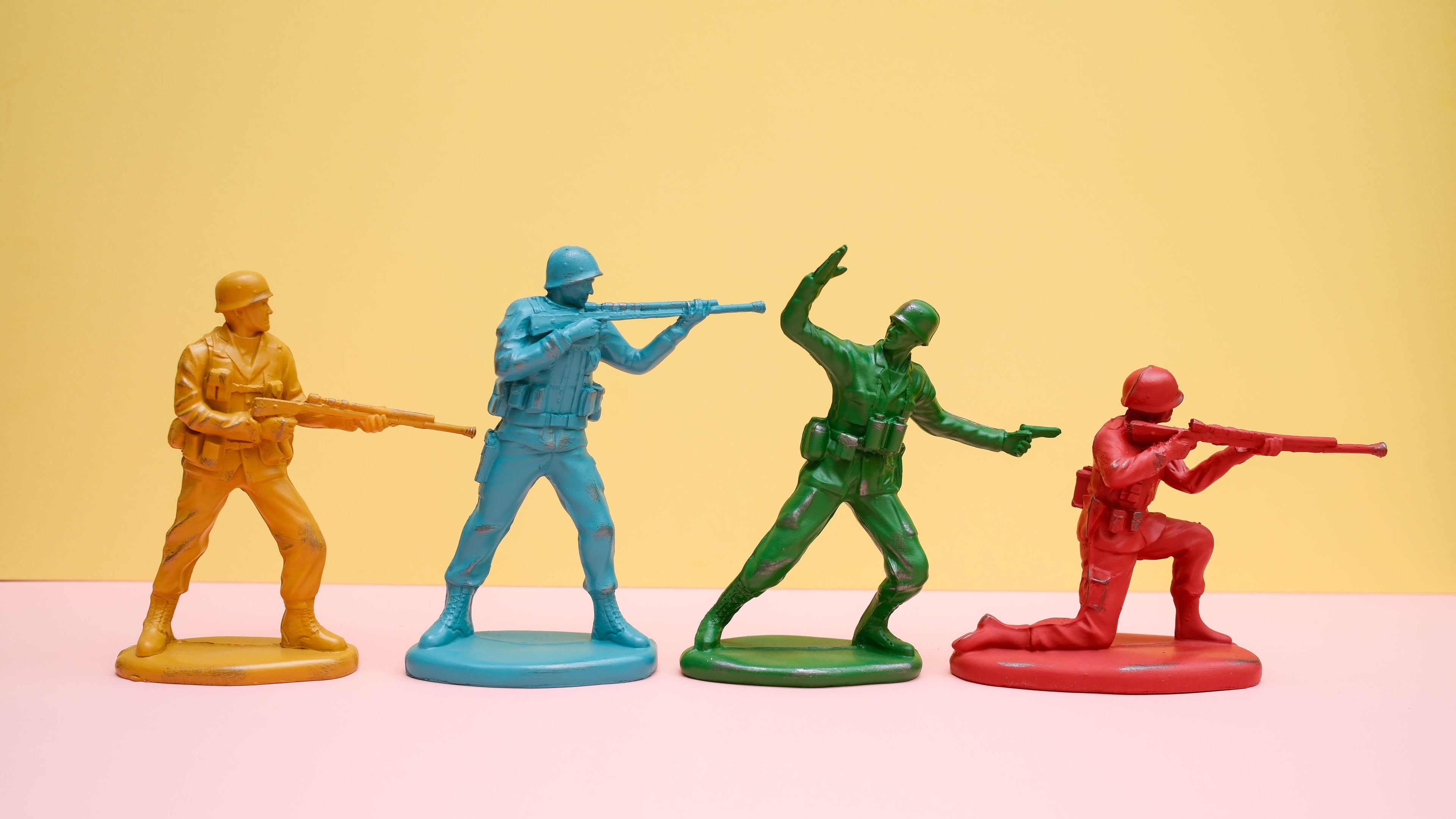Four toy soldier ornaments n different colors with weapons on a pink and yellow background