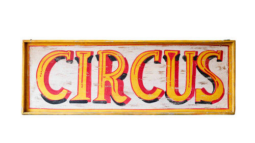 Wooden sign with 'CIRCUS' in red and yellow on a floral tablecloth against a wooden wall.