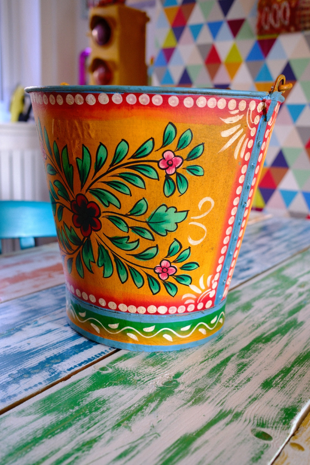 Colorful floral hand painted bucket on a table with a multicolored geometric patterned wall in the background