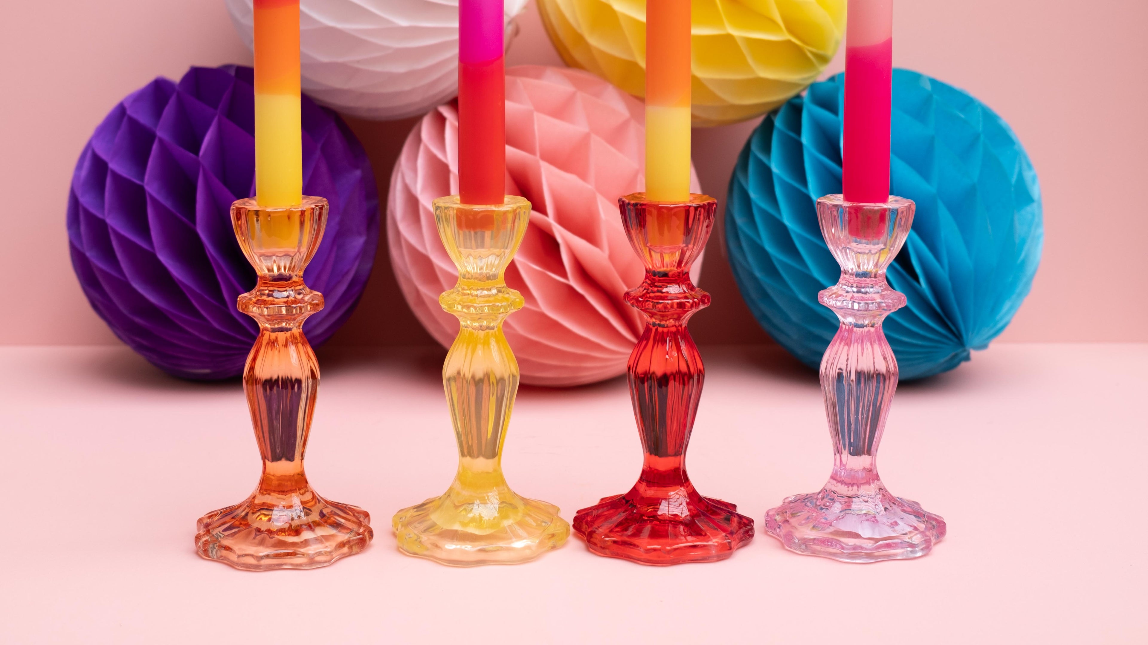 Colorful glass candlesticks with candles in front of colorful paper balls on a pink background