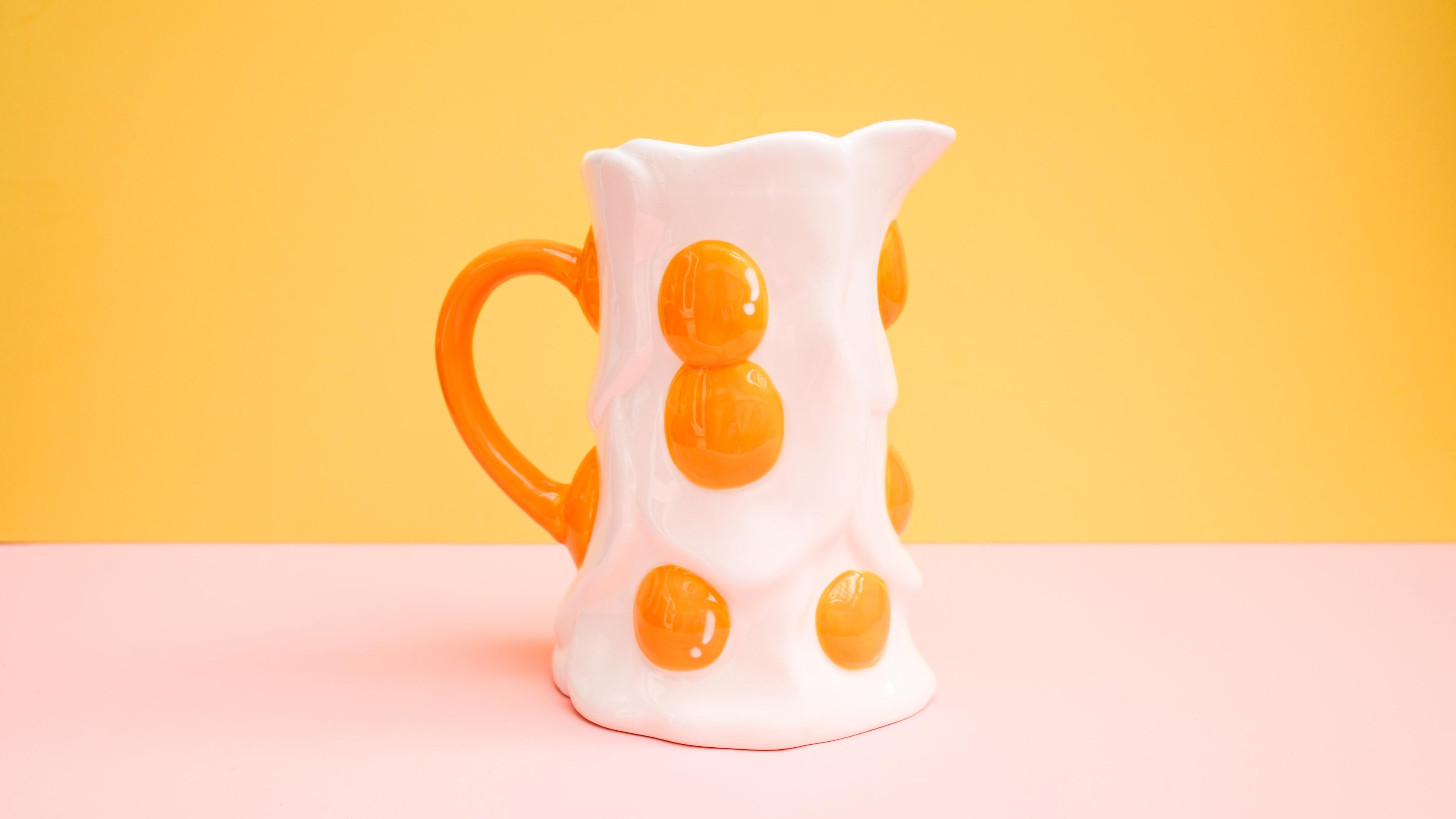 White ceramic pitcher with orange polka dots on a pink and yellow gradient background
