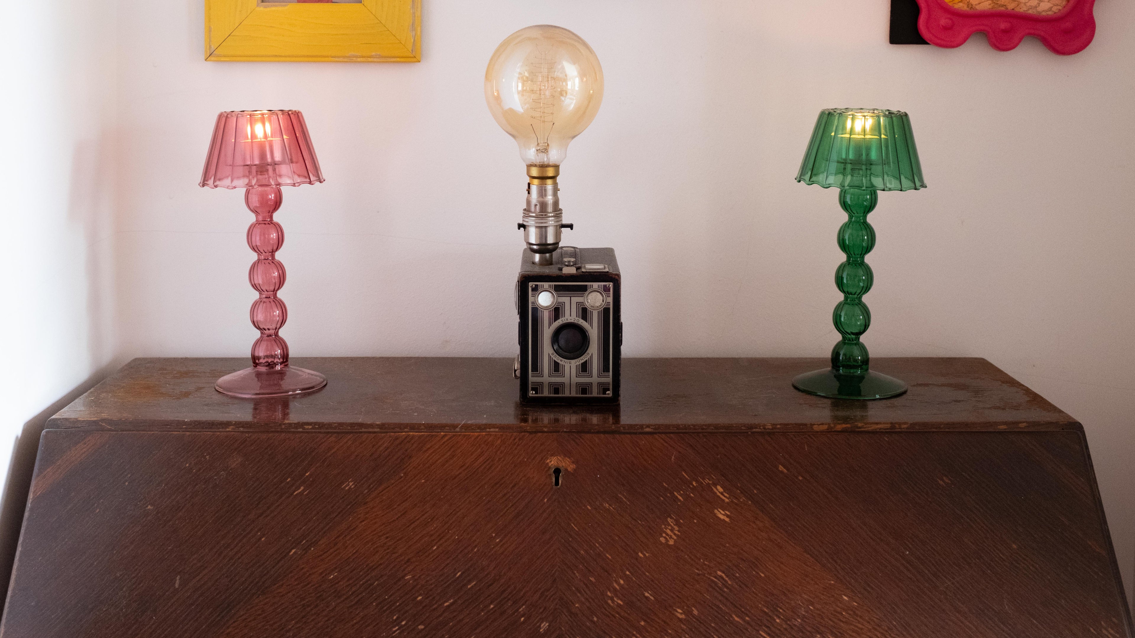 Three colorful lamps on a wooden table with framed pictures in the background.
