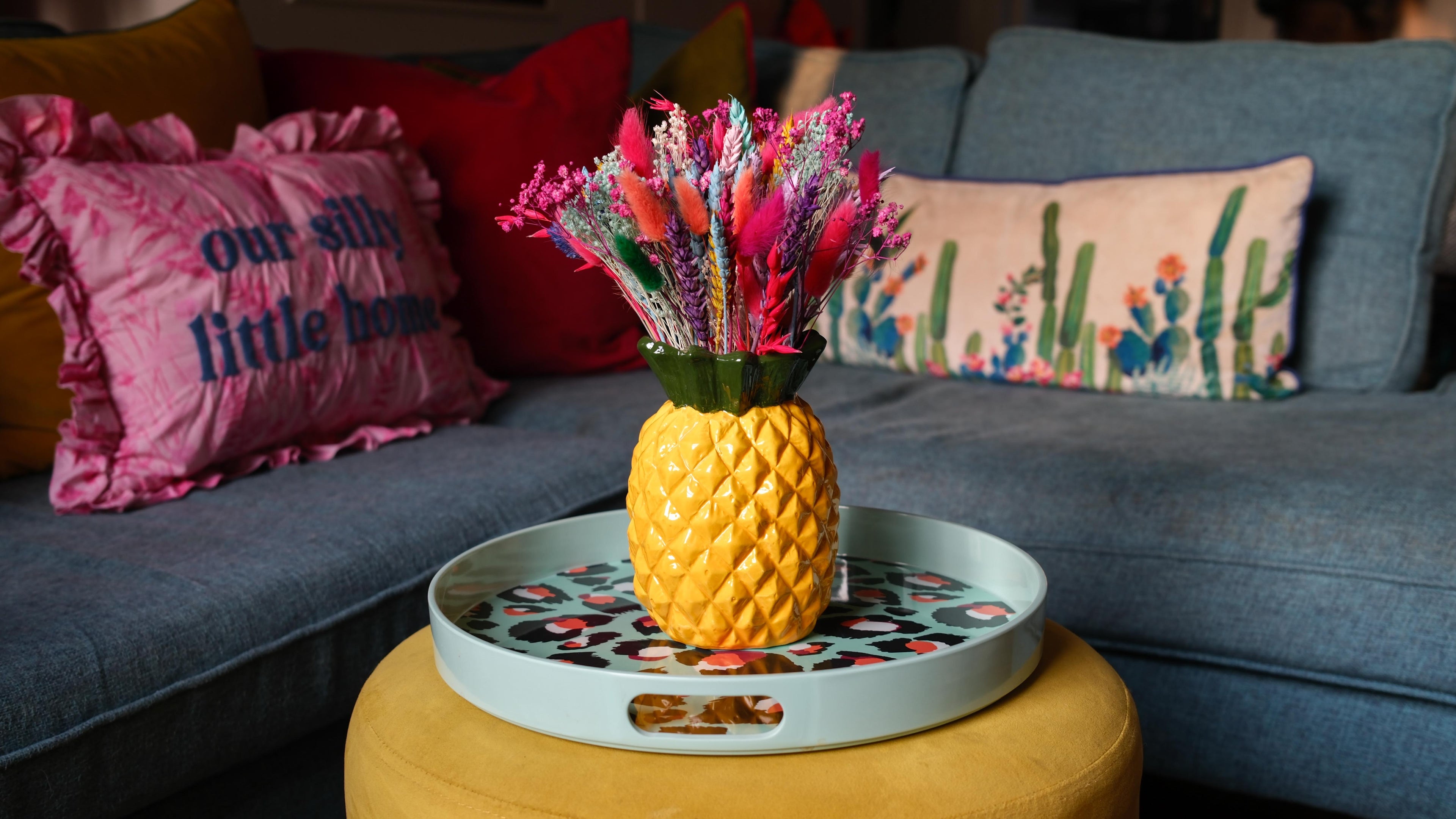 Yellow pineapple-shaped vase with pink flowers on a round tray in front of a gray couch with colorful pillows.