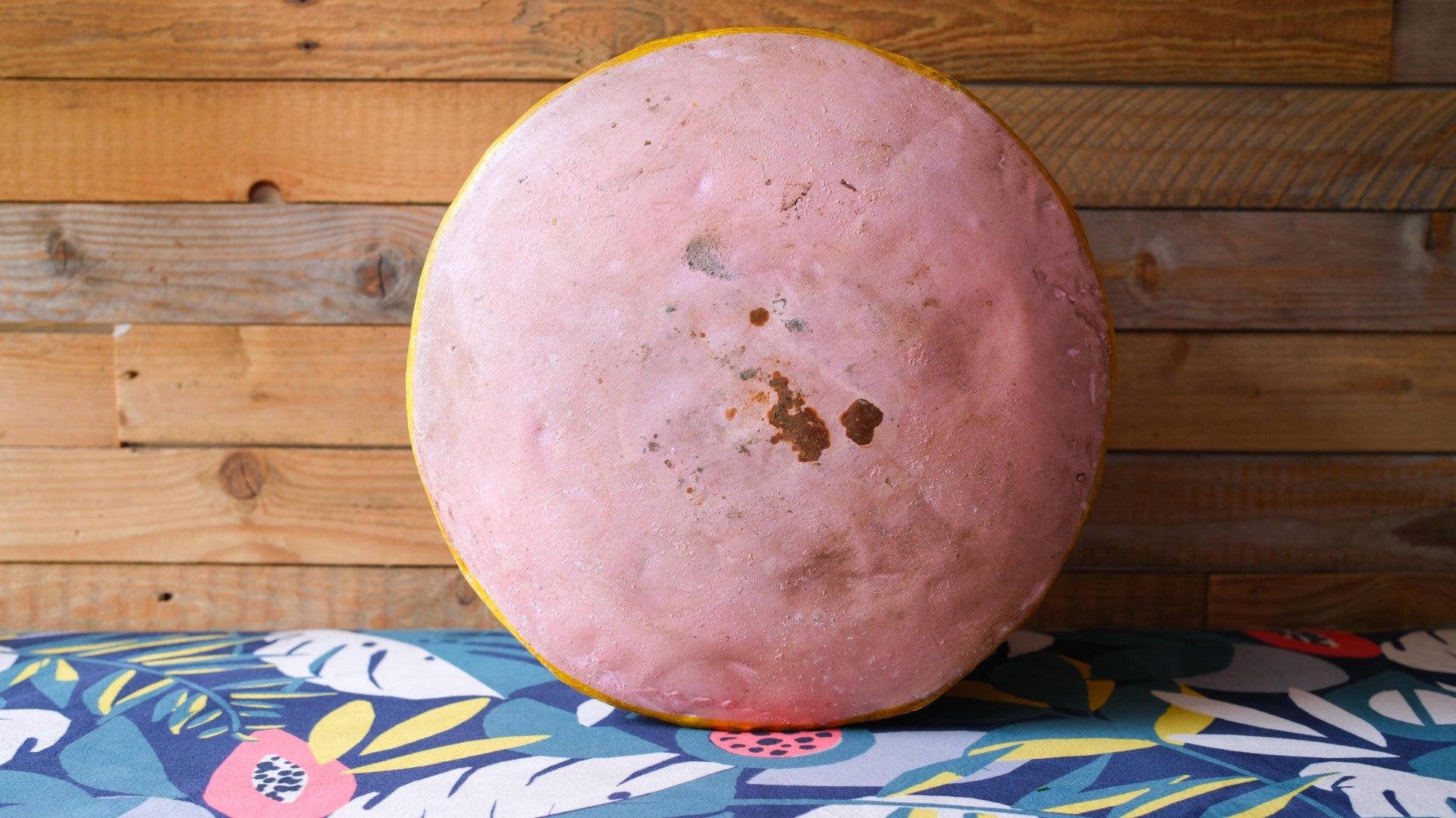 Round pink object with yellow rim on a colorful tablecloth against a wooden wall.
