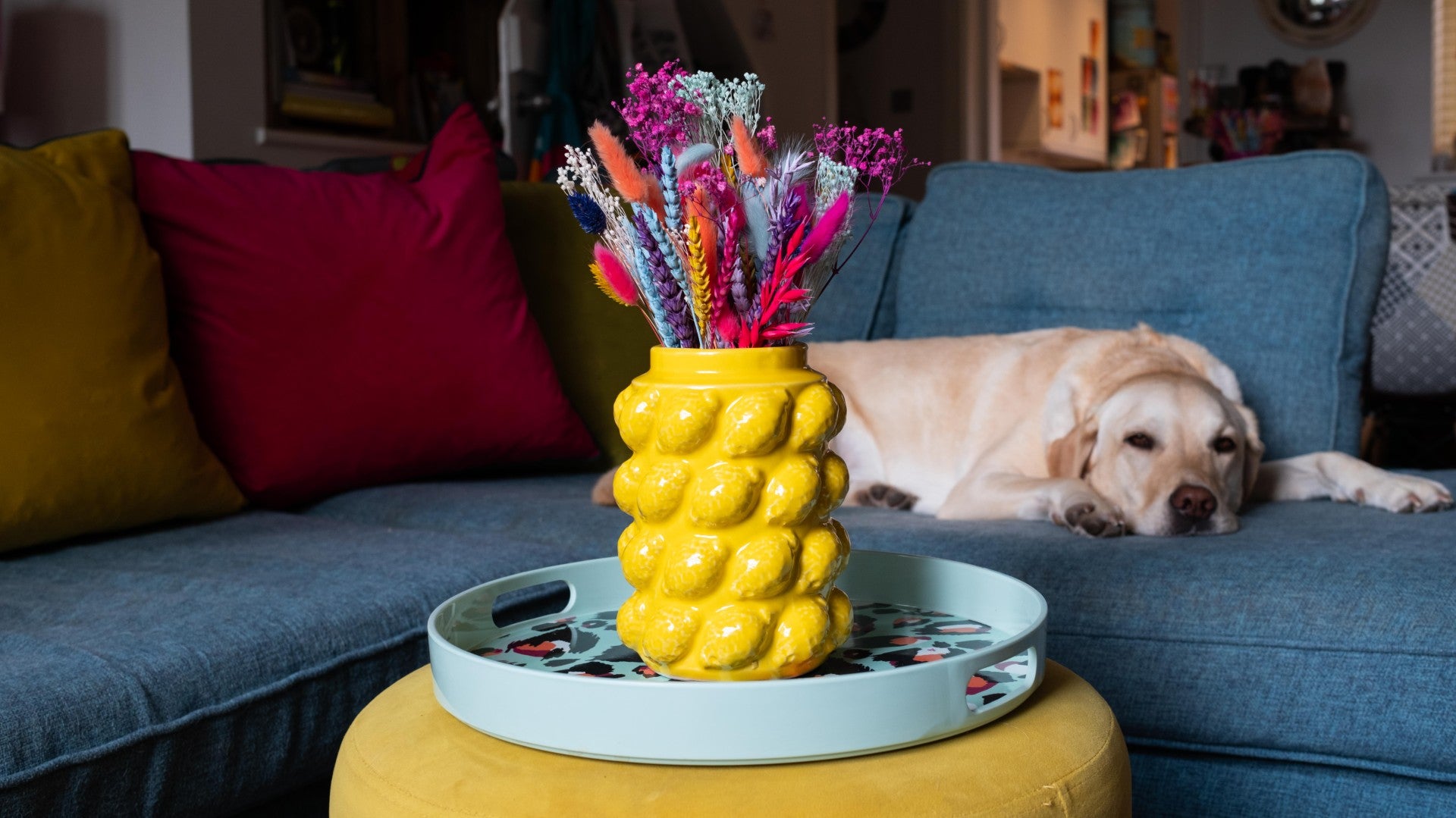 Yellow textured vase with colorful flowers on a tray, dog lying on a blue couch in the background