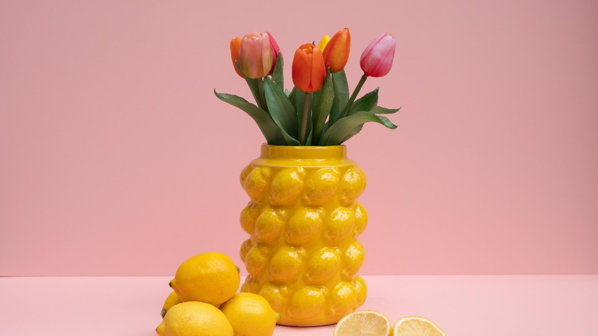 Yellow lemon shaped vase with tulips and lemons on a pink background