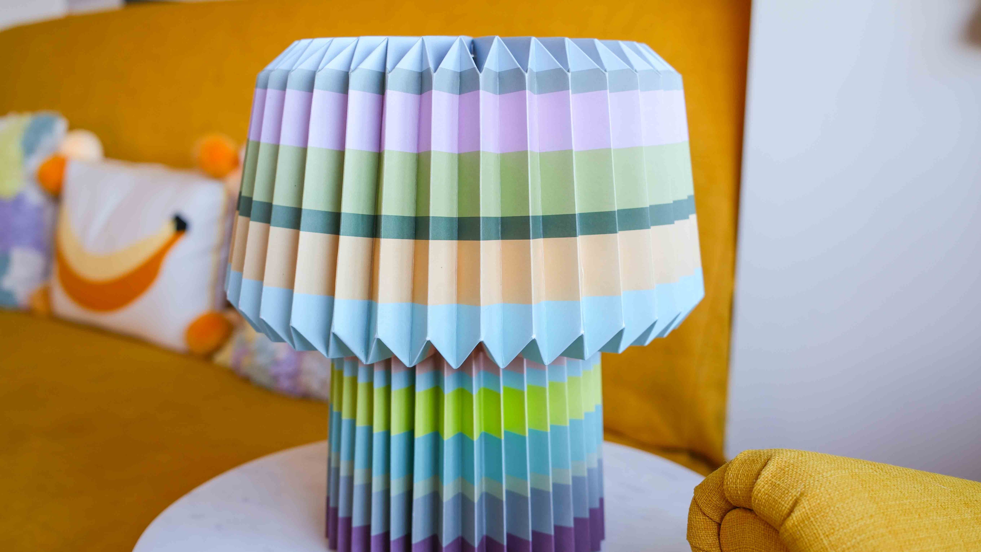 Colorful origami lamp on a white surface with a yellow couch and colorful pillow in the background
