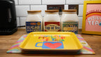Yellow tray with a colorful design on a kitchen counter with tea canisters in the background.