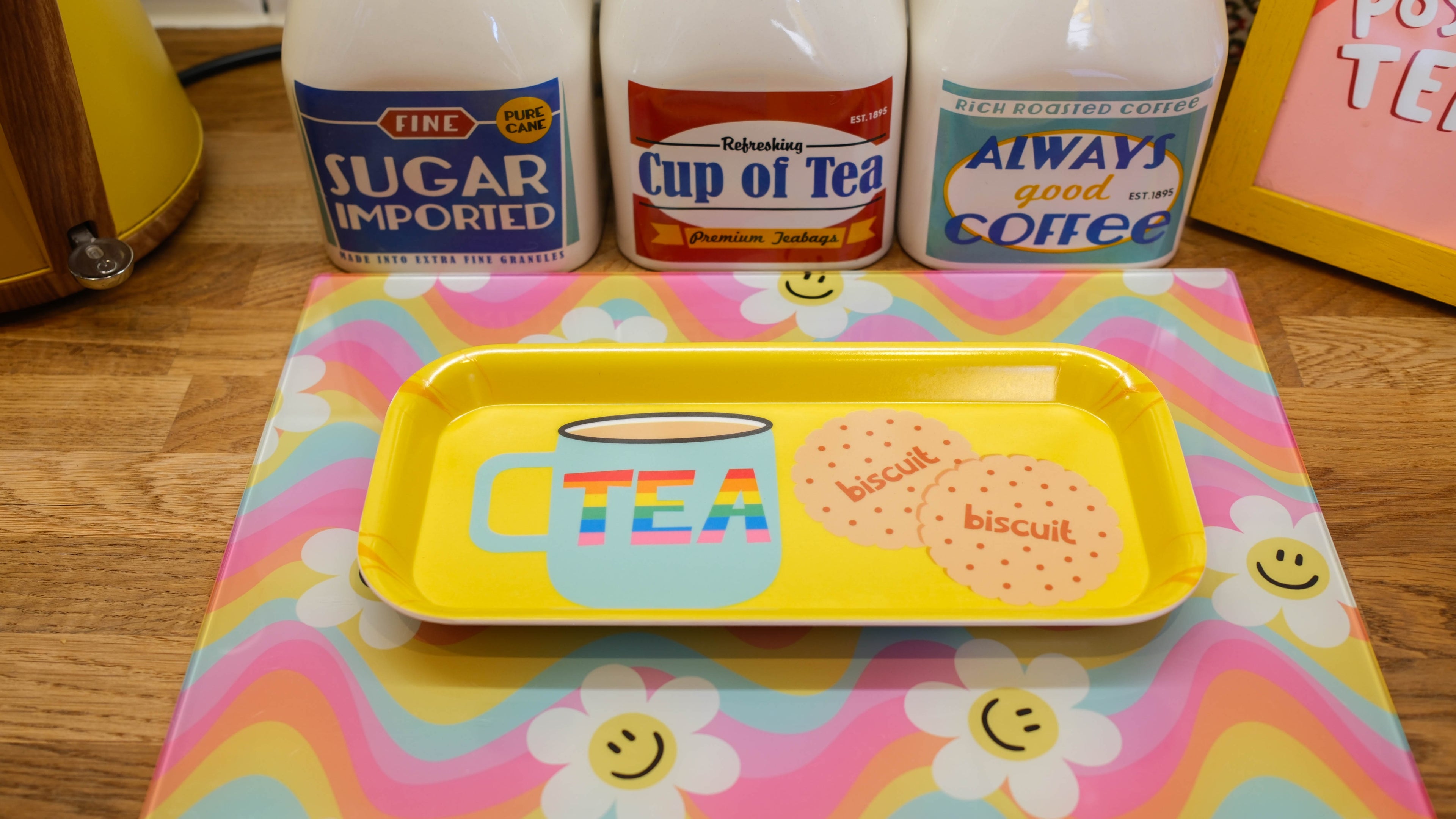 Yellow tray with 'Cup of Tea' design on a colorful placemat with tea-related products in the background.