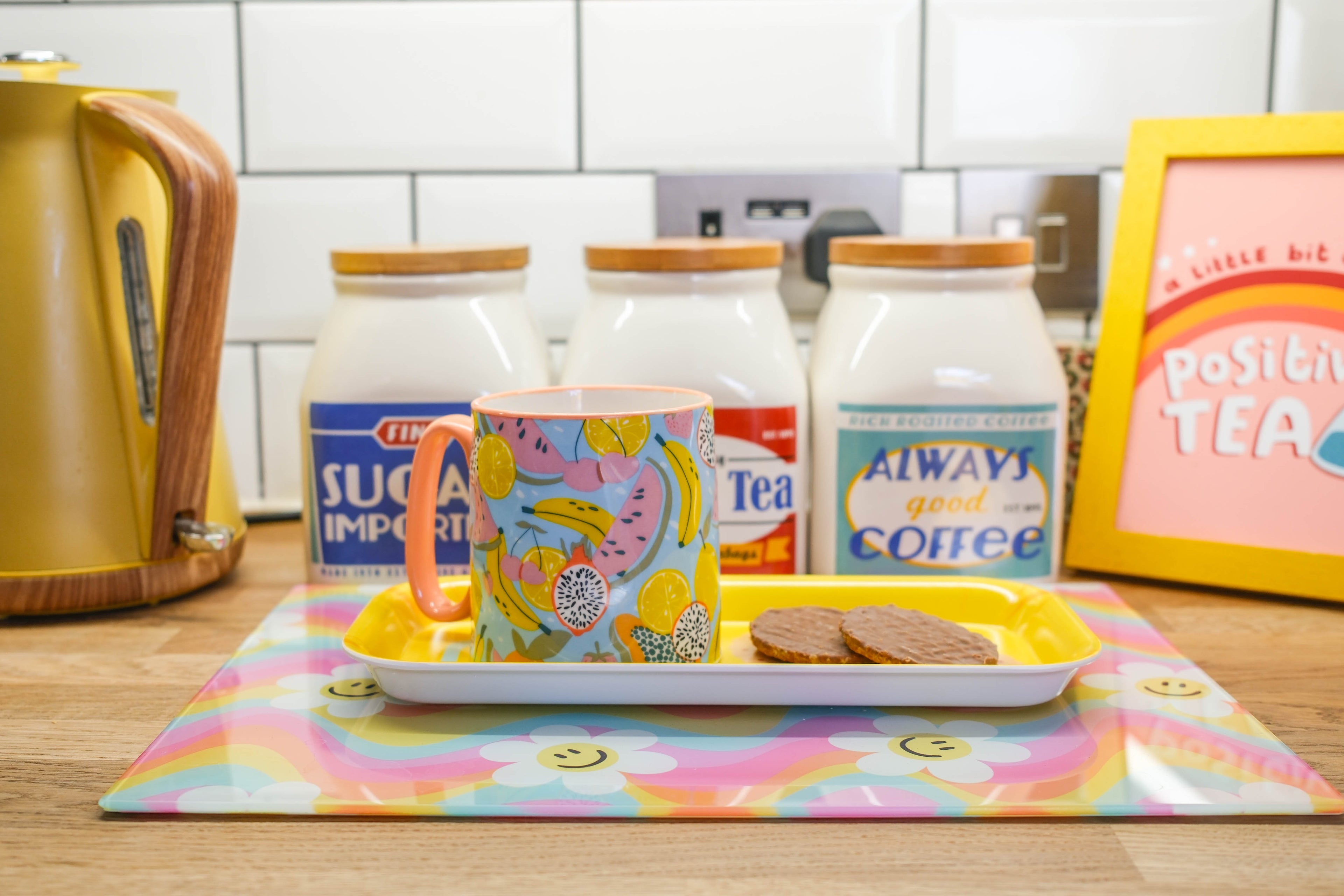 Colorful mug on a tray with cookies, surrounded by tea and coffee containers in a kitchen setting.