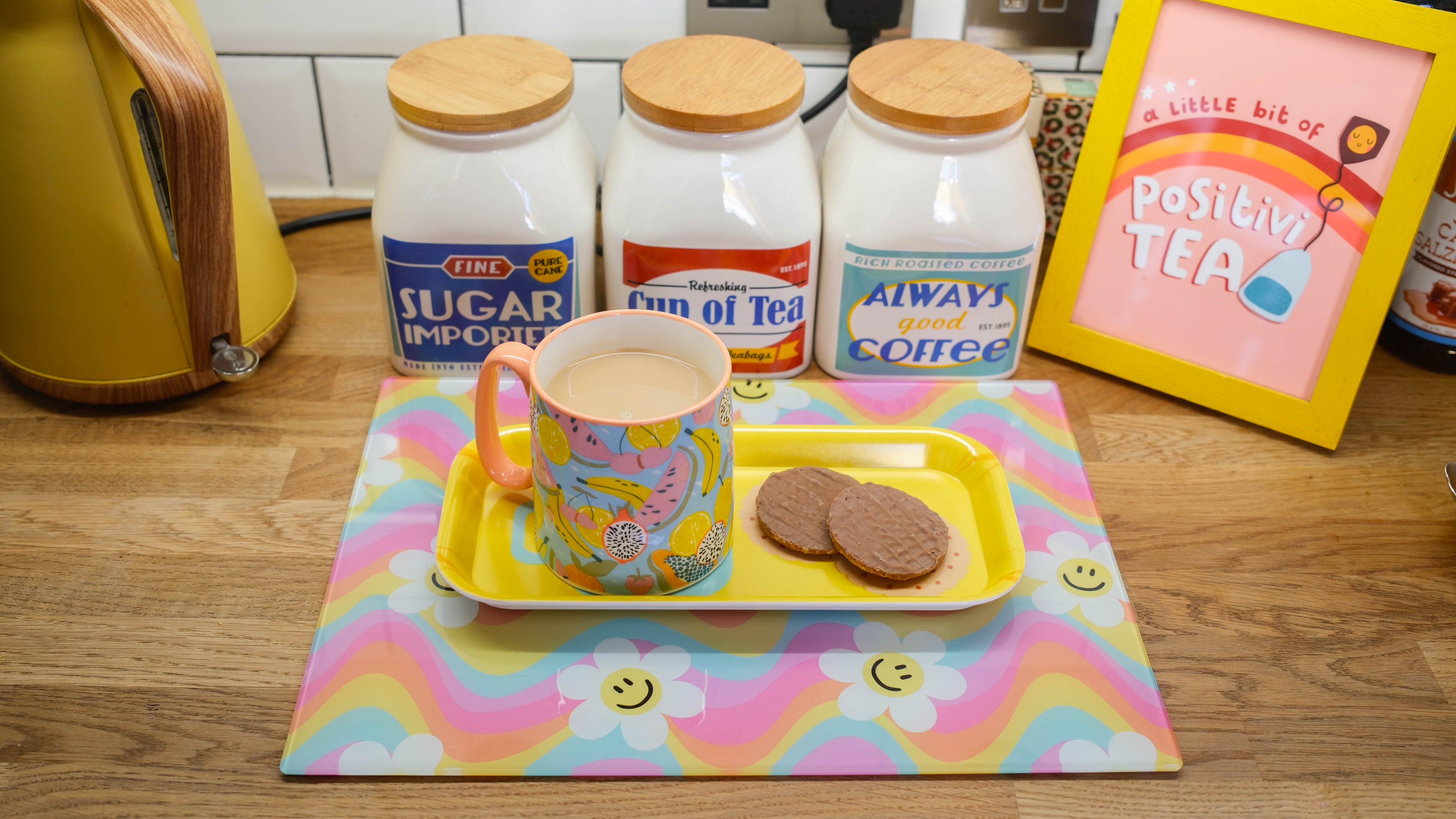 Cup of tea with biscuits on a tray, surrounded by jars of sugar, tea, and coffee on a kitchen counter.
